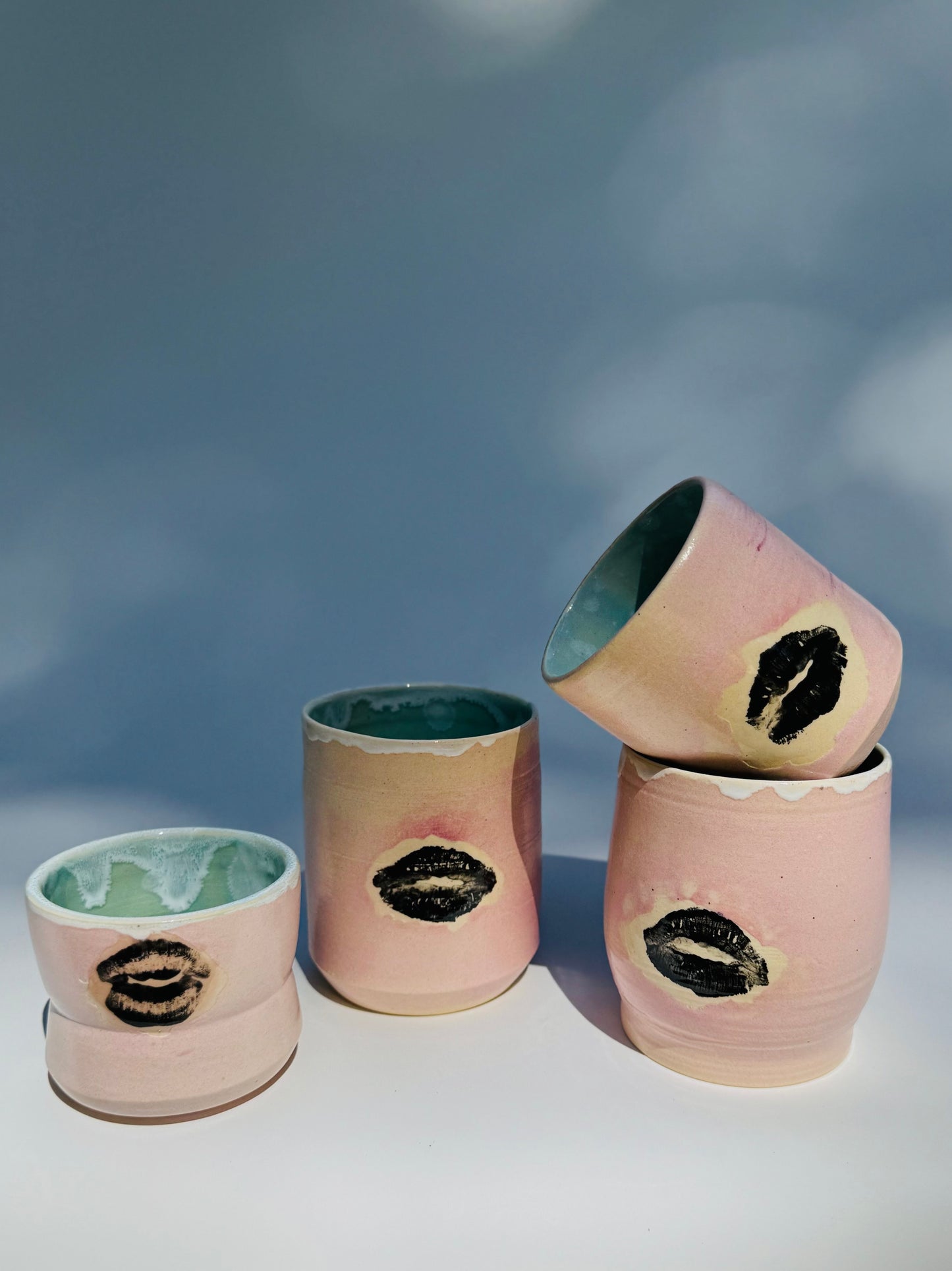 Set of four ceramic Kissy Cup Tumblers by Rita Strayer Ceramics of Savannah. This design features a cascading flux rim, various sea foam interior and celadon bloom shads. Stoneware, microwavable, black lip design with pink dominant colors and a blueish interior hue. Available in a variety of sizes.