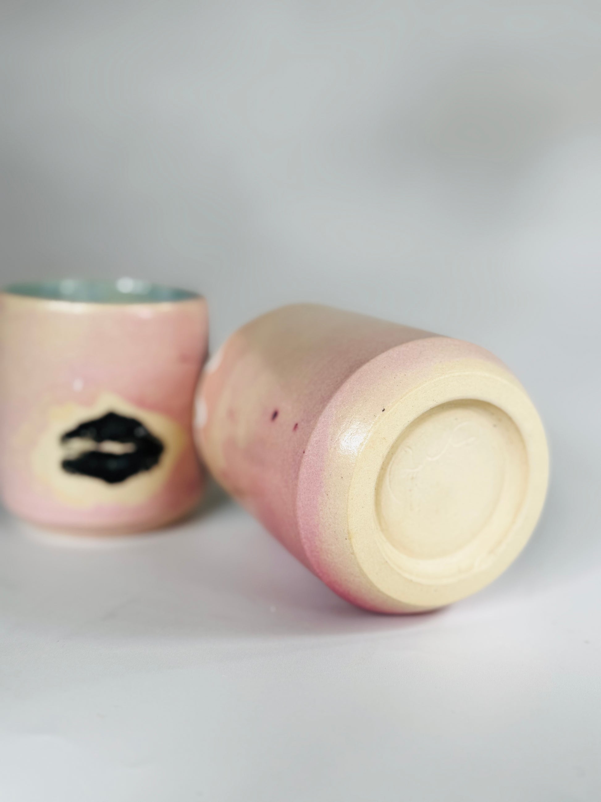 Rear-facing side view of the Large Kissy Cup Tumbler by Rita Strayer Ceramics of Savannah. Cascading flux rim, various sea foam interior and celadon bloom shads. Stoneware, microwavable, black lip design with pink dominant colors.