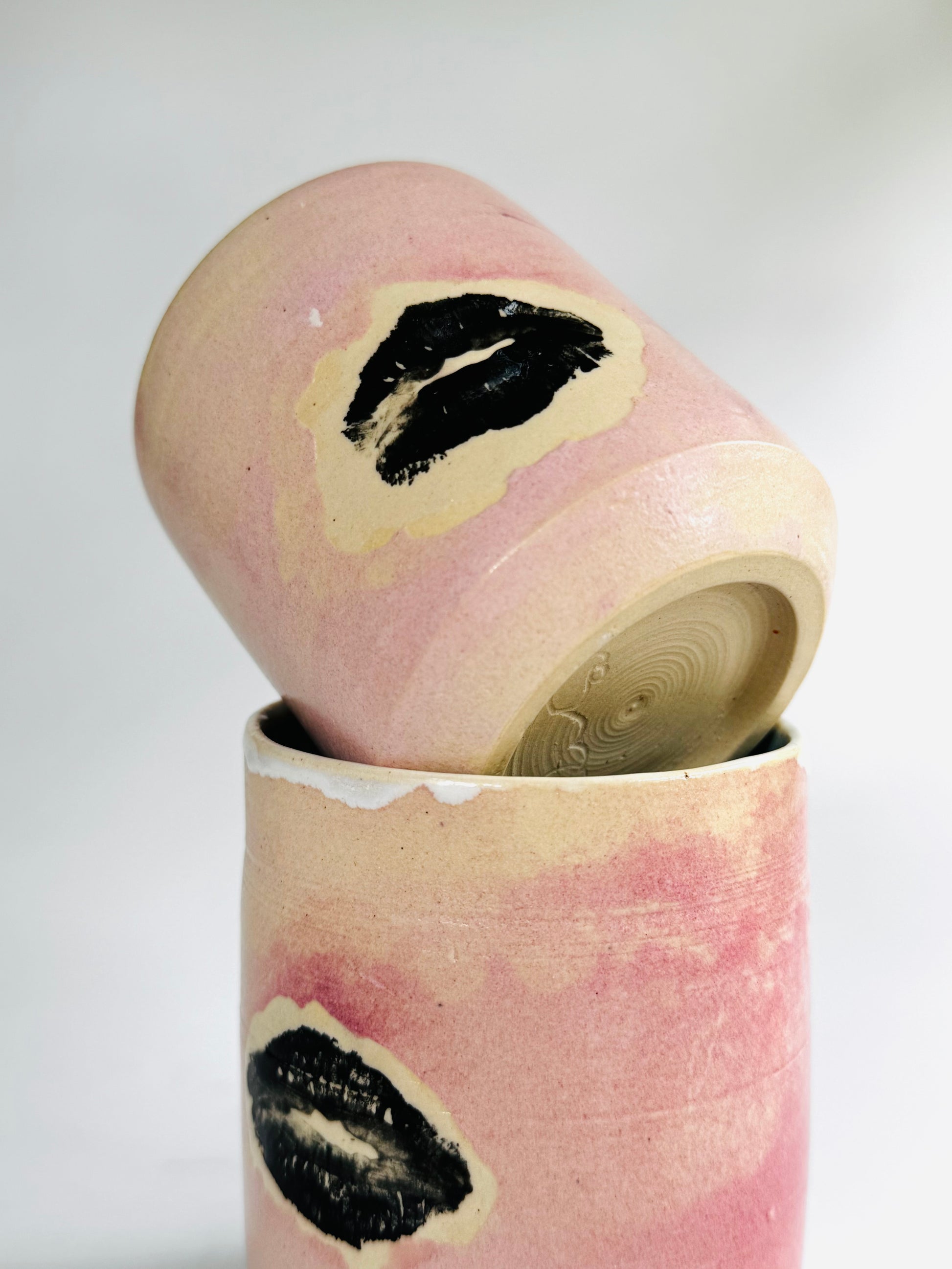 Playful view of two Small Kissy Cup Tumblers by Rita Strayer Ceramics of Savannah. This unique design features a cascading flux rim, various sea foam interior and celadon bloom shads. Stoneware, microwavable, black lip design with pink dominant colors and a blueish-hued interior.