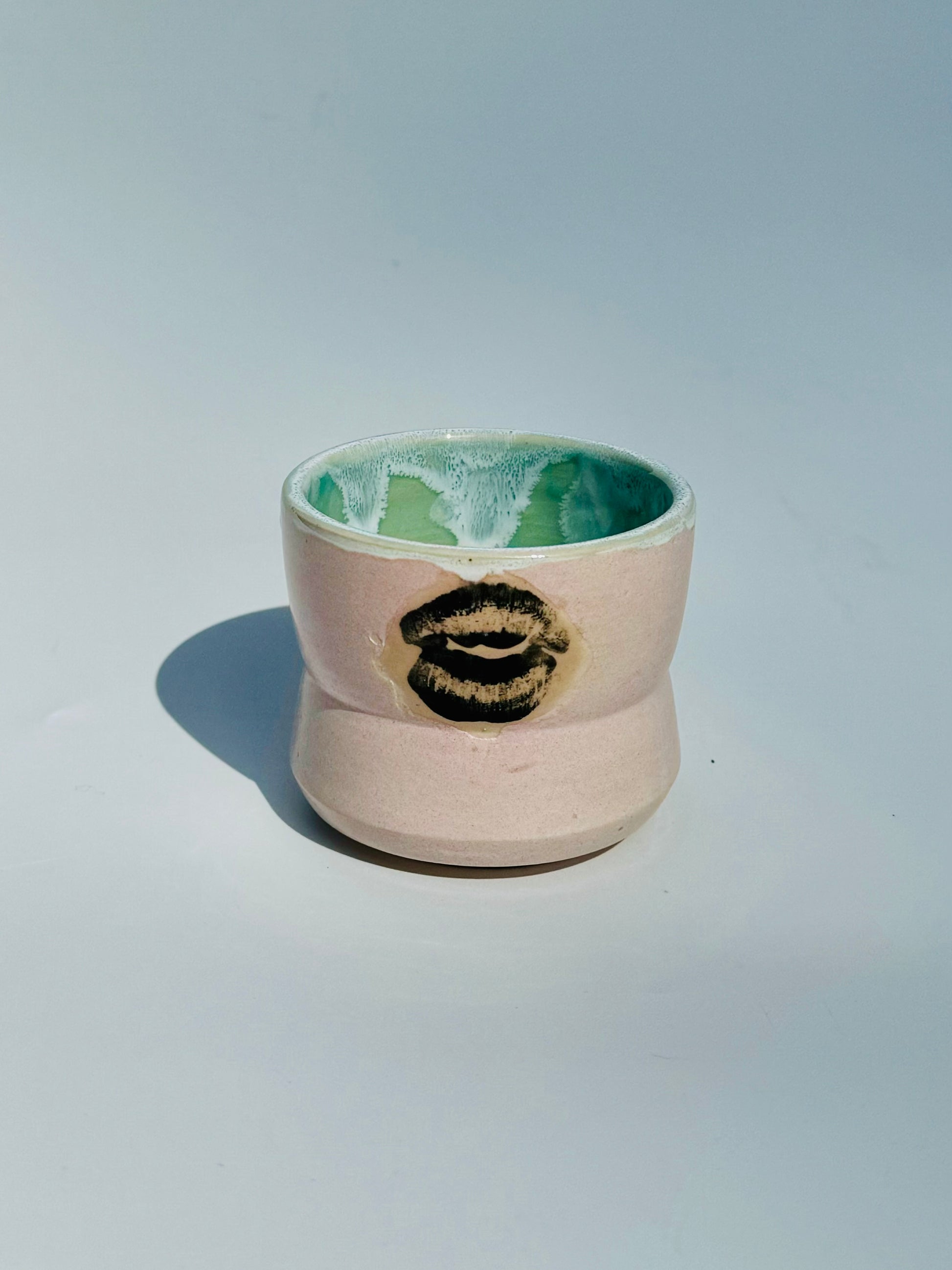 A straight ahead, slightly overhead view of the Kissy Cup Mini-sized ceramic cup. Created by Rita Strayer Ceramics of Savannah. Features green interior and black lip design on a light grey background.