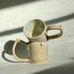 Teardrop Handle Mugs (pair) - only from Rita Strayer Ceramics of Savannah. Asymmetrical pair of mugs with a roomy handle hold. 8 oz Teardrop Handle Mugs via potters wheel, carved, & hand-built. Glazes dipped, sprayed, & hand-painted. View of two stacked mugs.