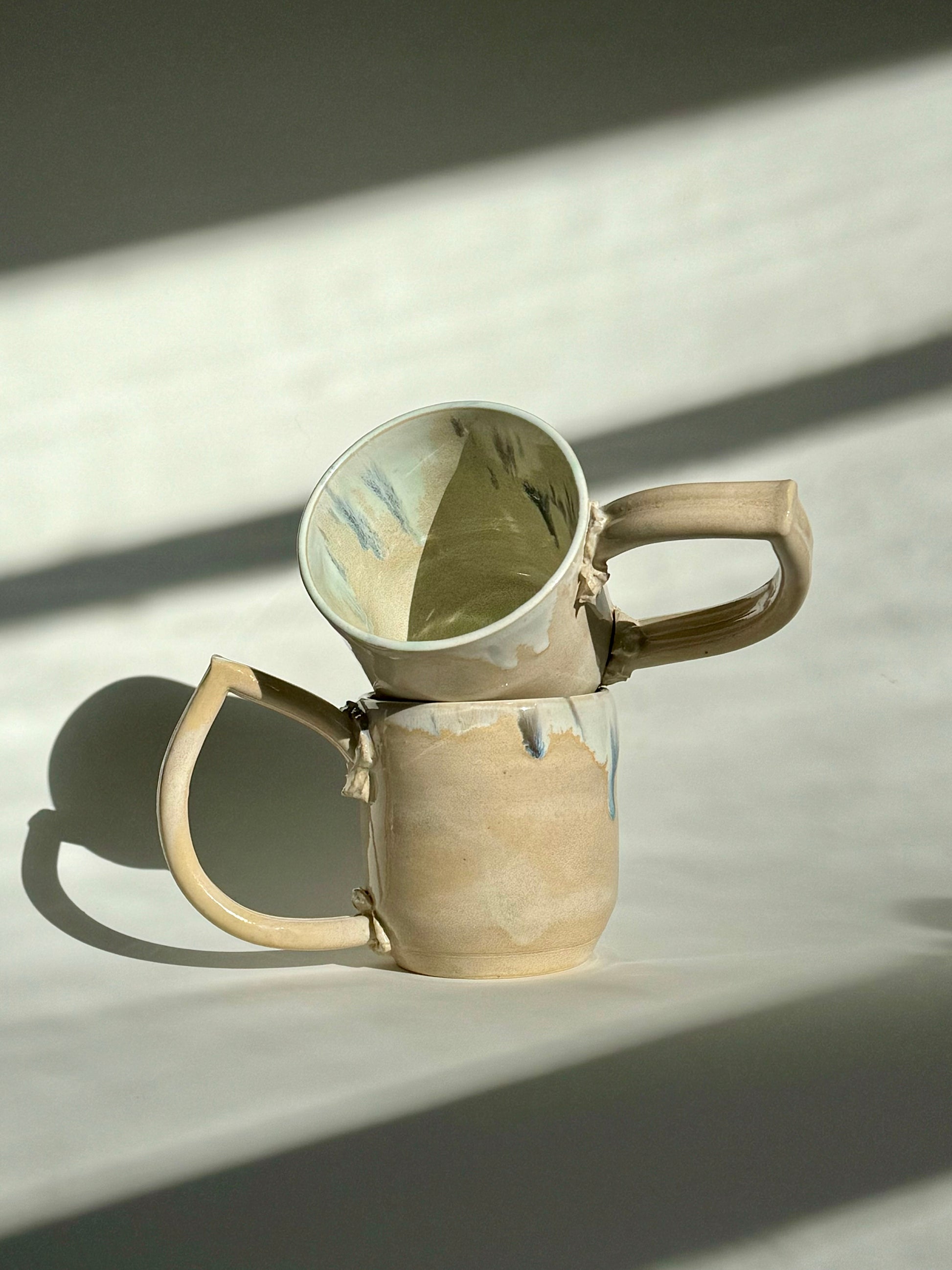Teardrop Handle Mugs (pair) - only from Rita Strayer Ceramics of Savannah. Asymmetrical pair of mugs with a roomy handle hold. 8 oz Teardrop Handle Mugs via potters wheel, carved, & hand-built. Glazes dipped, sprayed, & hand-painted. View of two stacked mugs.