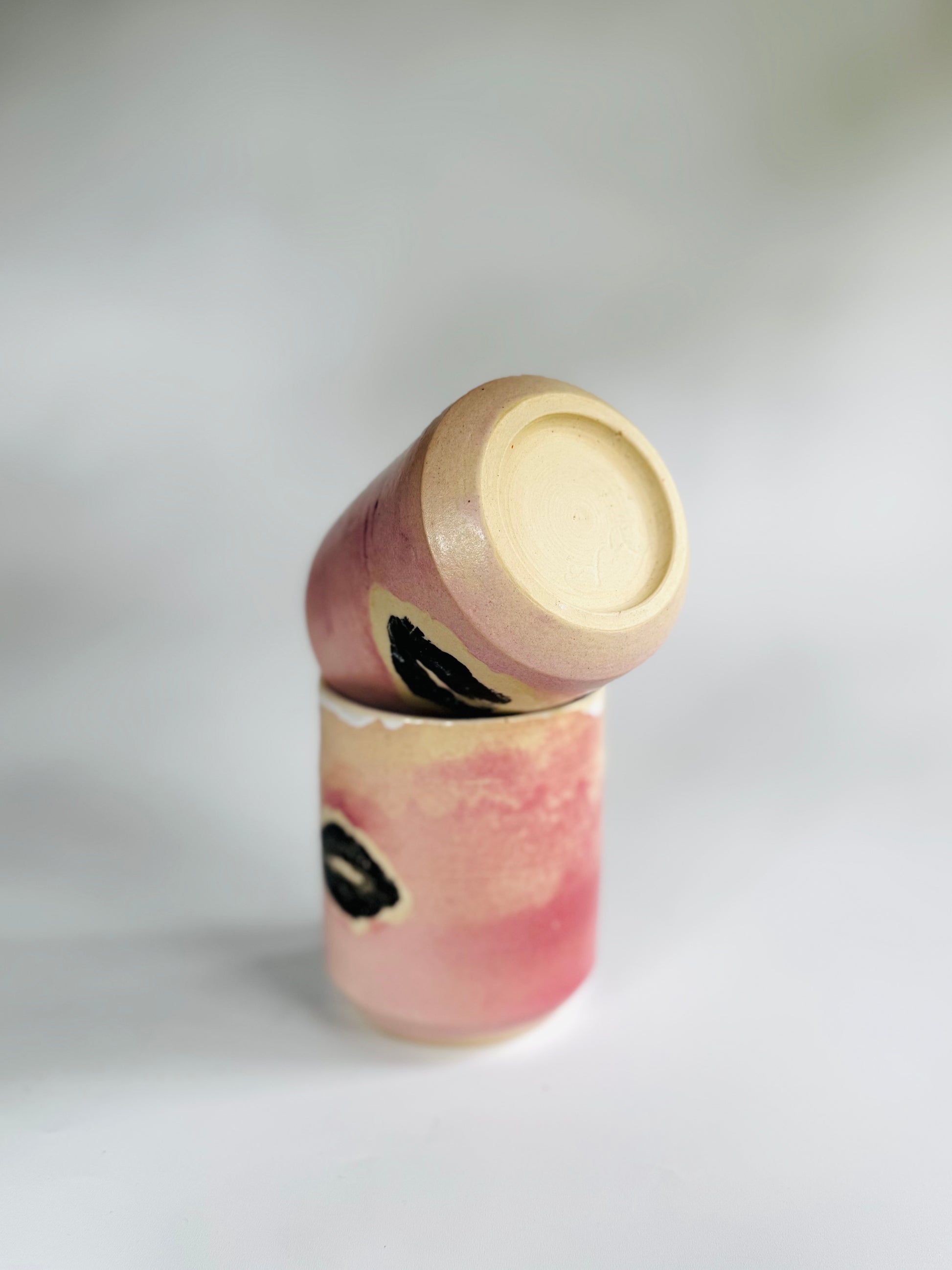 Another playful view of two Small Kissy Cup Tumblers by Rita Strayer Ceramics of Savannah. This unique design features a cascading flux rim, various sea foam interior and celadon bloom shads. Stoneware, microwavable, black lip design with pink dominant colors and a blueish-hued interior.