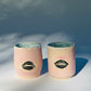 Distant exterior view of two Large Kissy Cup Tumblers by Rita Strayer Ceramics of Savannah. This tumbler features Cascading flux rim, various sea foam interior and celadon bloom shads. Stoneware, microwavable, black lip design with pink dominant colors.