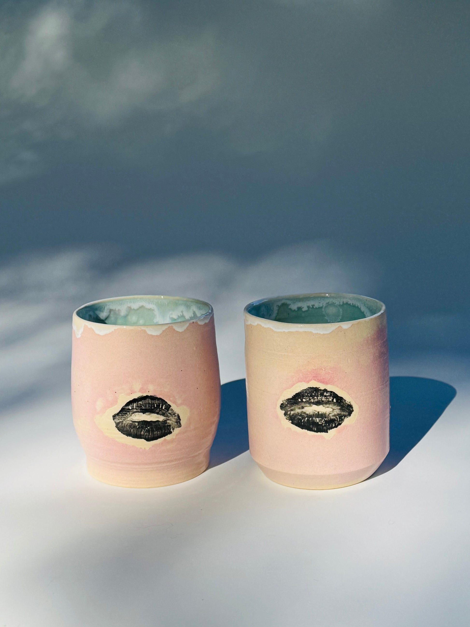 Distant exterior view of two Large Kissy Cup Tumblers by Rita Strayer Ceramics of Savannah. This tumbler features Cascading flux rim, various sea foam interior and celadon bloom shads. Stoneware, microwavable, black lip design with pink dominant colors.