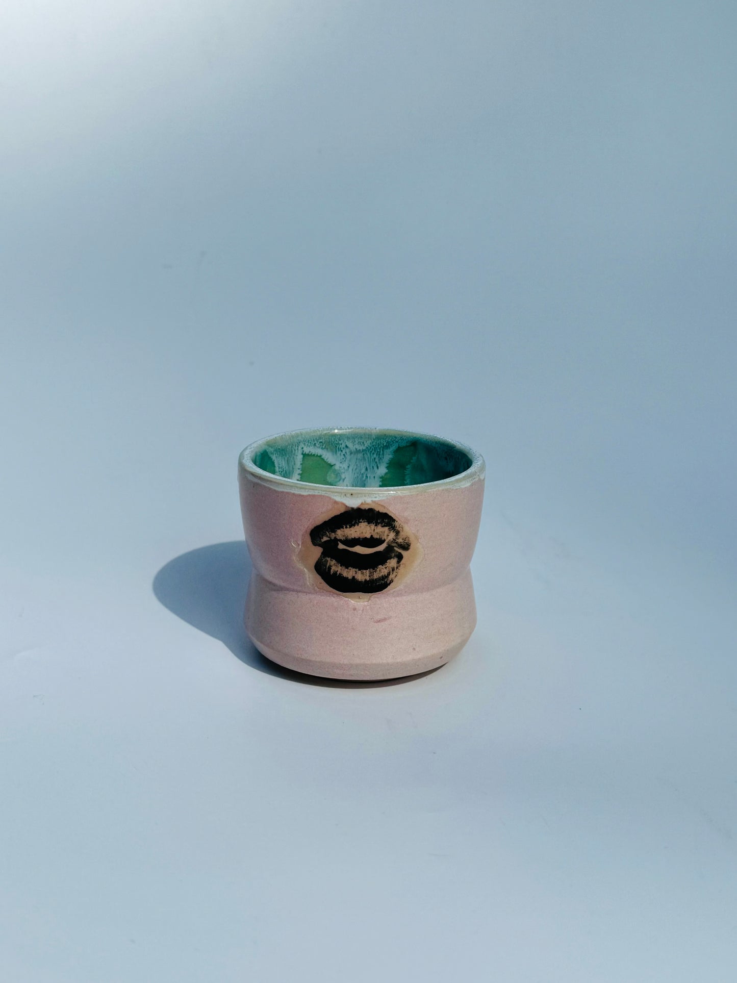 Distant view of the Kissy Cup Mini-sized ceramic cup. Created by Rita Strayer Ceramics of Savannah. Features green interior and black lip design on a light grey background.
