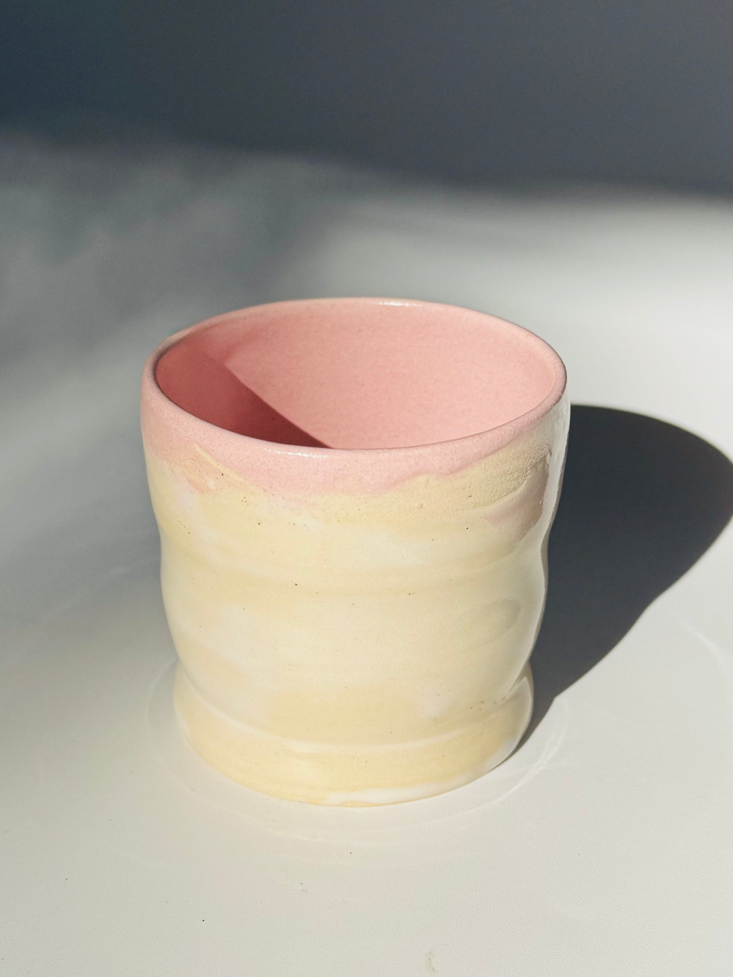Slightly overhead view of the Little Milk Cup tumbler design by Rita Strayer Ceramics of Savannah, GA. A tumbler with convenient finger grooves to accommodate little hands, sleepy hands, and slippery hands too. Two-tone pink and beige colouring.