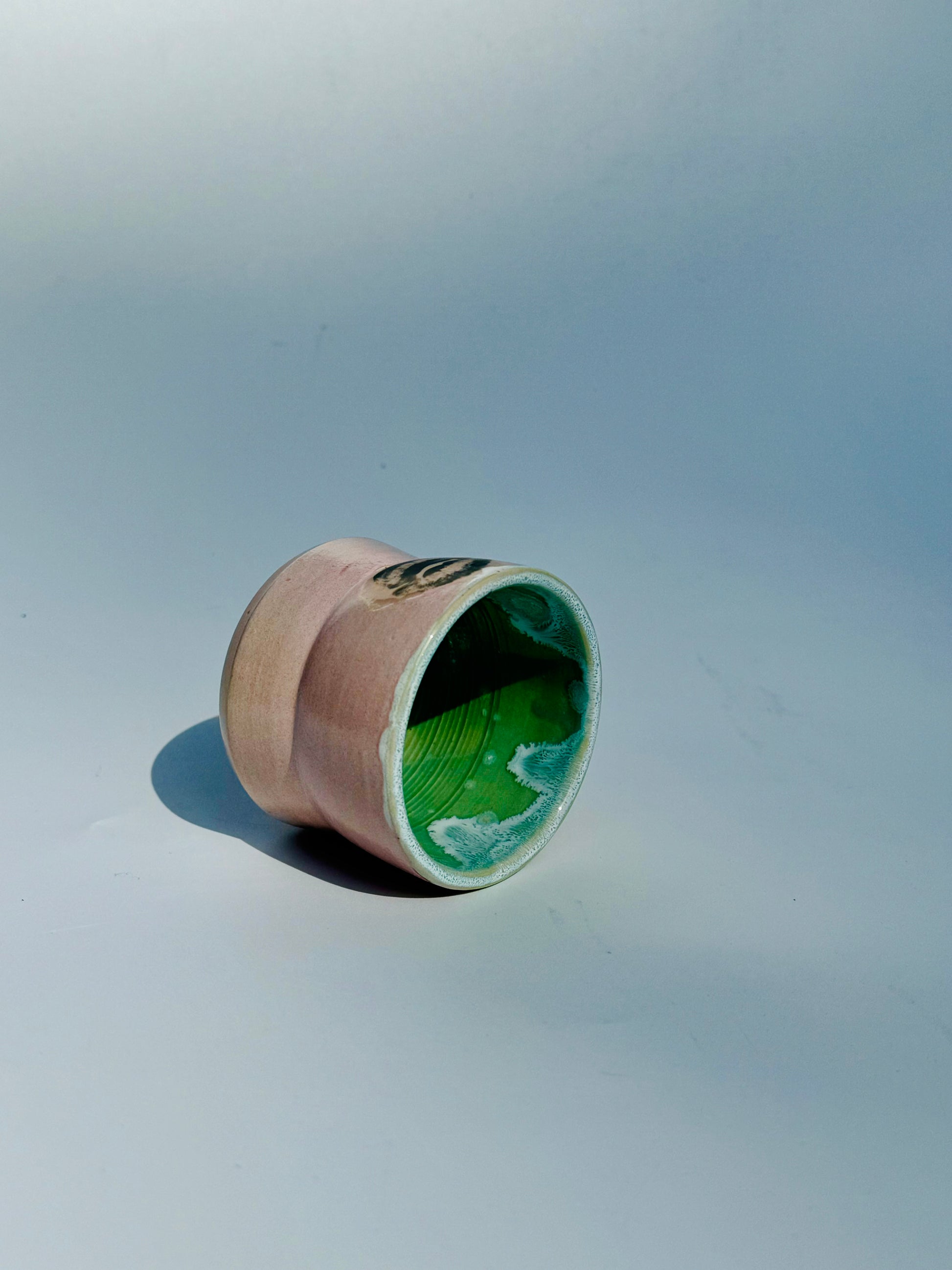 Tipped on its side view of the Kissy Cup Mini-sized ceramic cup. Created by Rita Strayer Ceramics of Savannah. Features green interior and black lip design on a light grey background.